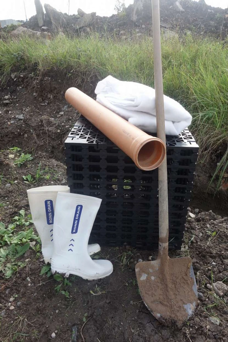 Nástroje pro odvodnění zahrady: lopata, boty, trubka a drenážní krabice.