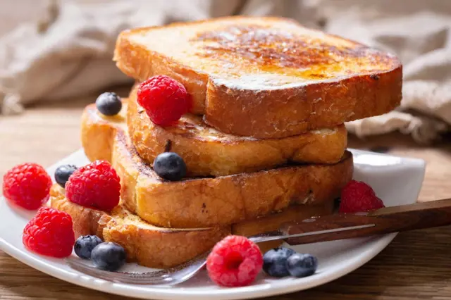 Zlatohnědý francouzský toasts s malinami a borůvkami na bílém talíři.