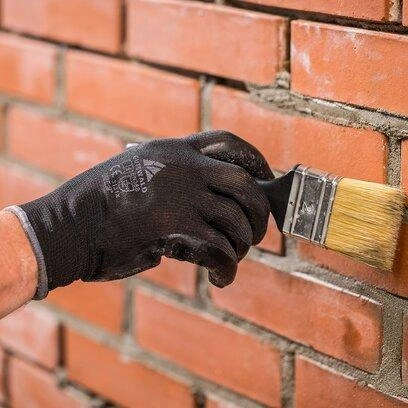 Ruka v rukavici nanáší maltu na režné zdivo štětcem.