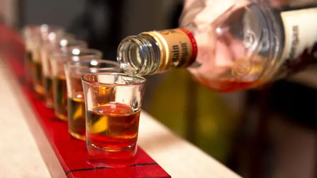 Nalévání alkoholu do skleniček na červeném podkladu.