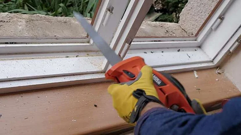 Člověk řezá bílé dřevěné okenné křídlo pilou při renovaci.
