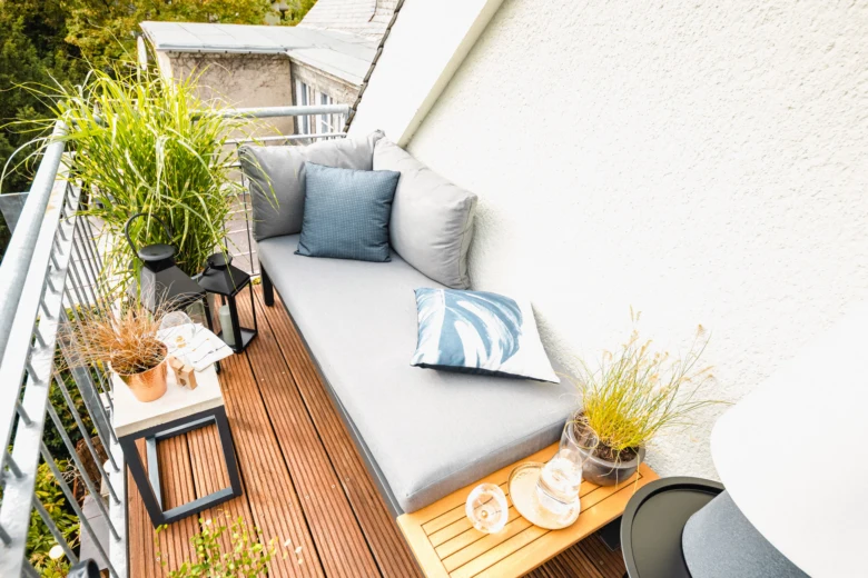 Zelený balkon s pohovkou, polštáři a rostlinami na dřevěné podlaze.