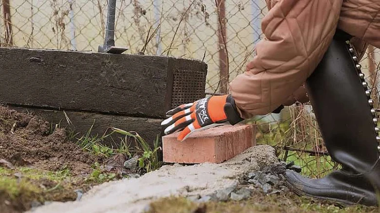 Ruka s rukavicí položí cihlu na zem vedle dřevěného rámu.