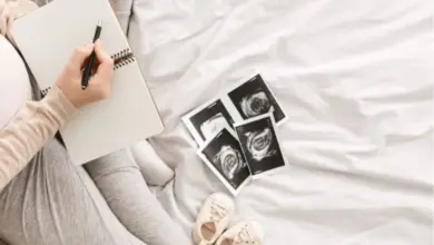 Těhotná žena píše do sešitu vedle ultrazvukových snímků a dětských botiček.