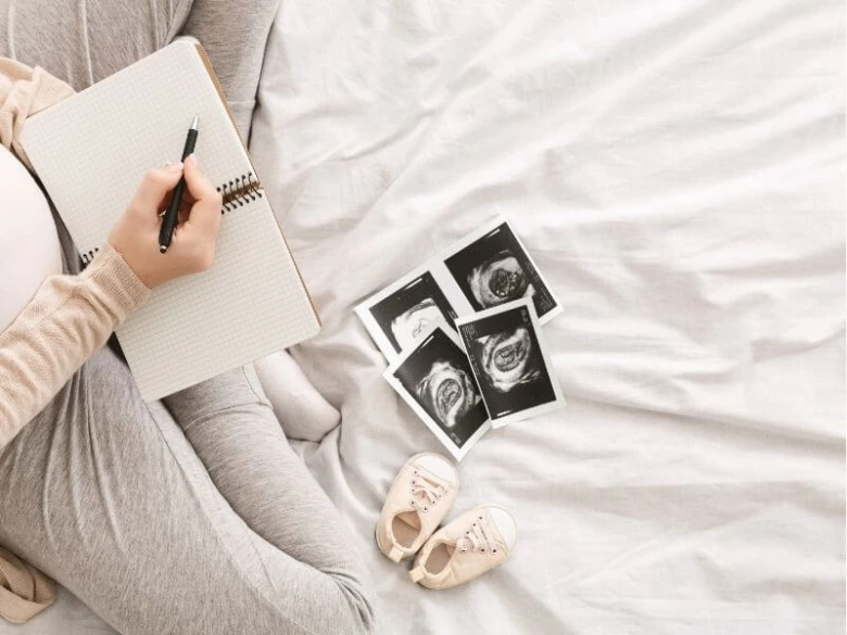 Těhotná žena píše do sešitu vedle ultrazvukových snímků a dětských botiček.